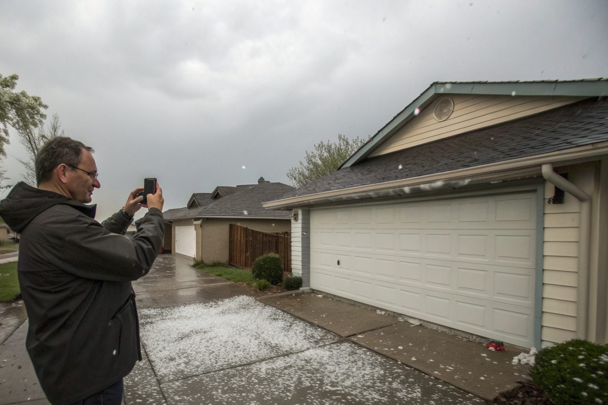 A man photographing his garage roof with a phone after a hailstorm, with hail on the driveway