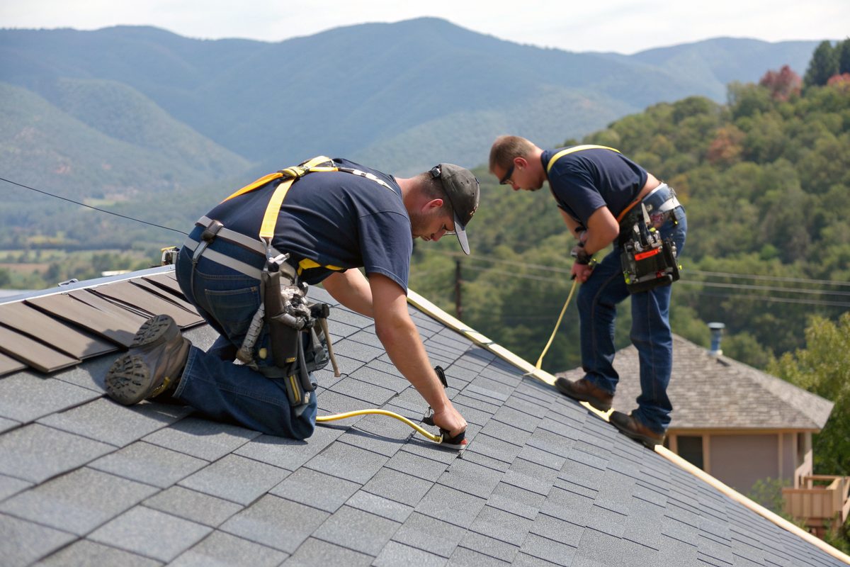 Two roofers in harnesses fastening new gray shingles with the foothills behind them
